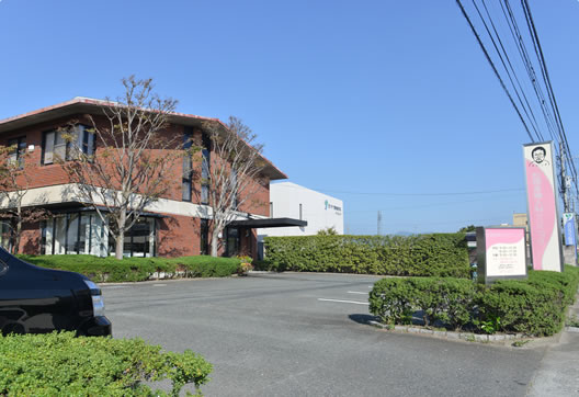 写真：松隈産婦人科クリニック外観　煉瓦色の建物　周囲を植栽に囲まれている　道路沿いに白とピンクでデザインされた看板が二枚立っている