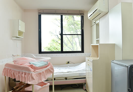 写真：病室の様子　奥の壁には大きな窓、左側の壁には棚、右側の壁にはエアコンがついていて、部屋の奥に母親用のベッド、手前に赤ちゃん用のベッドが置いてある。エアコンの下には荷物を入れられる大きな棚があり、その横には冷蔵庫がある。